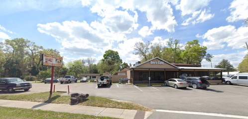 A restaurant with a sign, surrounded by parked cars and trees under a partly cloudy sky.