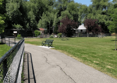 A park path leads to a grassy area with benches, trees, and a gazebo in the background on a sunny day.