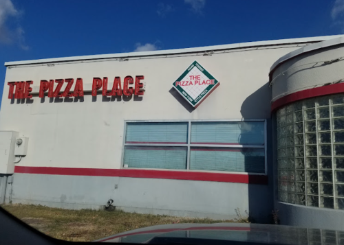 A building with a sign reading "The Pizza Place," featuring a glass block wall and blue sky in the background.