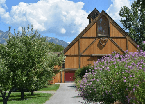 A wooden barn with a heart-shaped window, surrounded by trees and colorful flowers, under a blue sky with clouds.