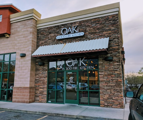 Exterior of Oak Wood Fire Kitchen, featuring a stone facade and large glass doors, with a sign above the entrance.