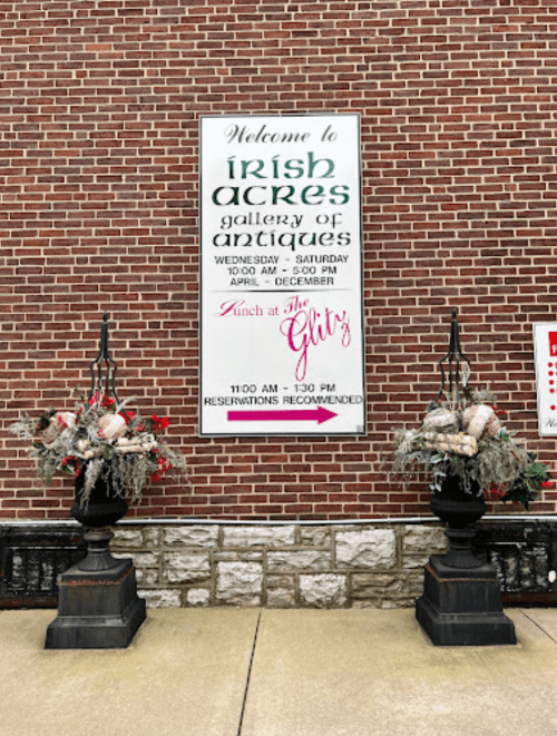 Sign for Irish Acres gallery of antiques, with floral arrangements on either side, set against a brick wall.