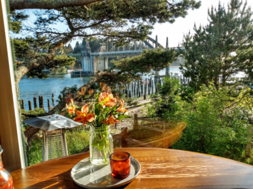 A scenic view from a table with a vase of flowers, overlooking a river and bridge surrounded by trees.