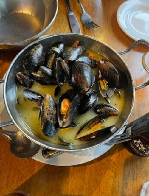 A metal bowl filled with mussels in a broth, accompanied by a spoon and a bottle on a wooden table.