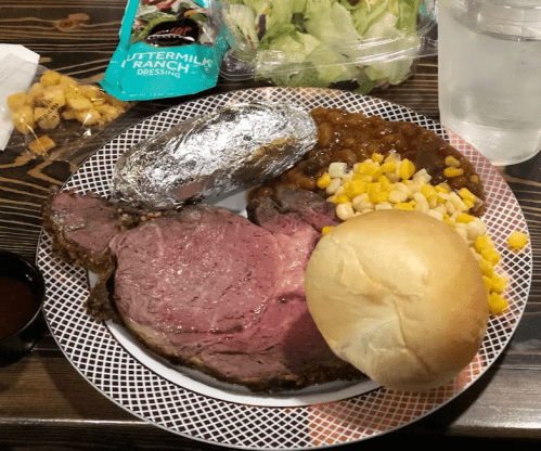 A plate with prime rib, baked potato, corn, baked beans, a roll, and a side salad with dressing.