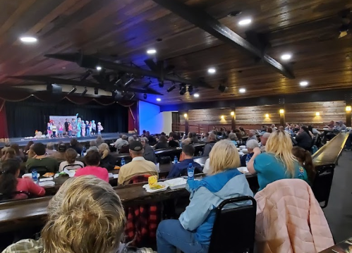 A crowded theater audience watches a performance on stage, with colorful costumes and a wooden interior.