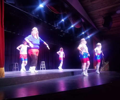 A group of performers in colorful workout outfits dancing on stage with bright lights and a black curtain backdrop.