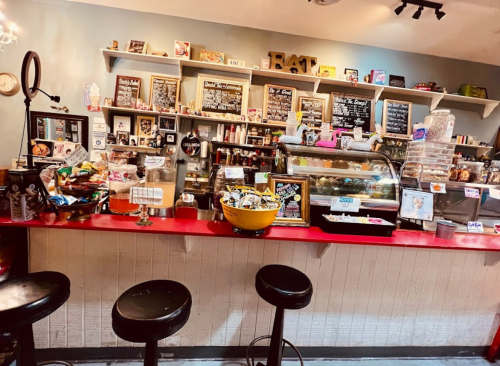 A cozy café interior featuring a red counter, stools, and a display of baked goods and snacks on shelves.