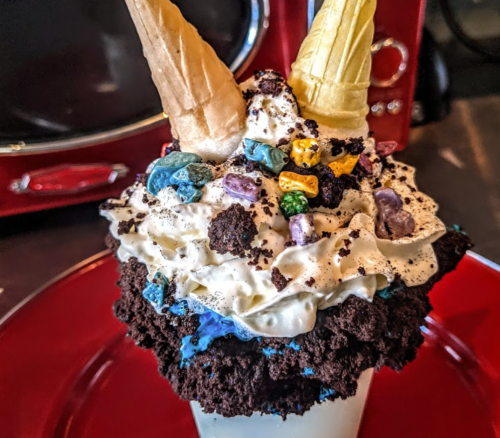 A colorful dessert with whipped cream, cookie crumbles, candy pieces, and two ice cream cones on top.