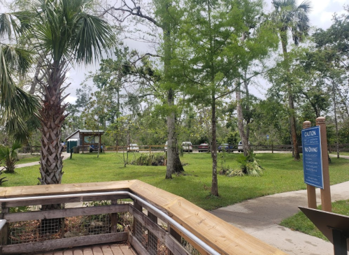 A wooden boardwalk leads to a green park with trees, a gazebo, and a sign indicating no fishing.
