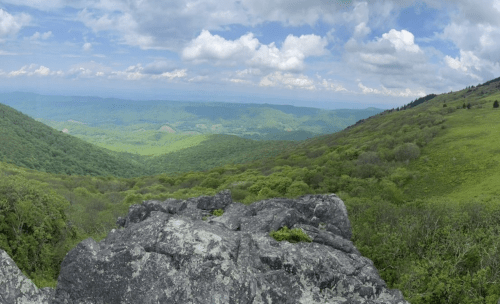 A panoramic view of lush green mountains under a partly cloudy sky, with rocky foreground and rolling hills in the distance.