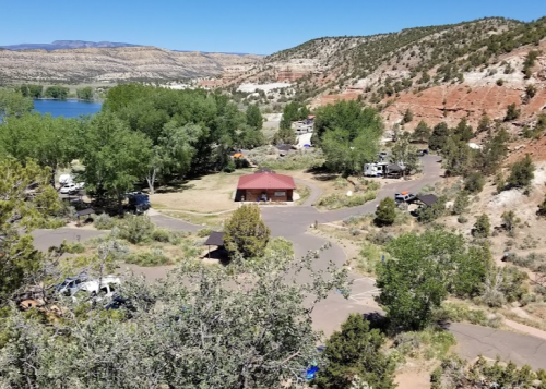 Aerial view of a campground with a red-roofed building, surrounded by trees and mountains under a clear blue sky.