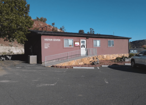 A visitor center building with a ramp, surrounded by a parking lot and trees in a rocky landscape.