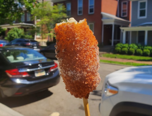 A close-up of a golden-brown corn dog on a stick, with a blurred street and houses in the background.