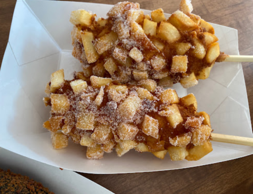 Two skewers of fried dough coated in cinnamon sugar and topped with diced apples, served in a white paper tray.