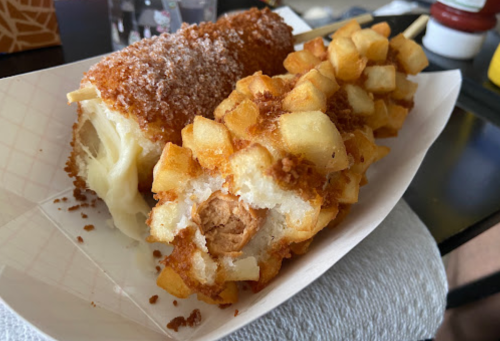 A close-up of a plate featuring a corn dog coated in breadcrumbs and a potato-covered sausage on a stick.