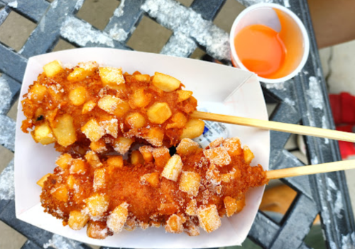 Two crispy corn dogs topped with diced potatoes, served with a small cup of orange sauce on a checkered table.