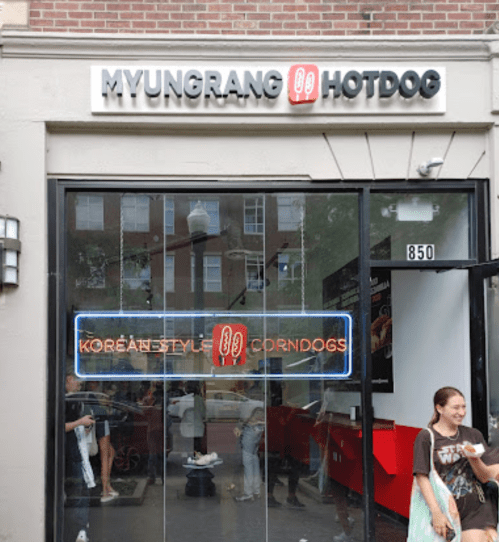 Exterior of Myungrang Hotdog, featuring a neon sign for Korean-style corn dogs, with people outside the entrance.