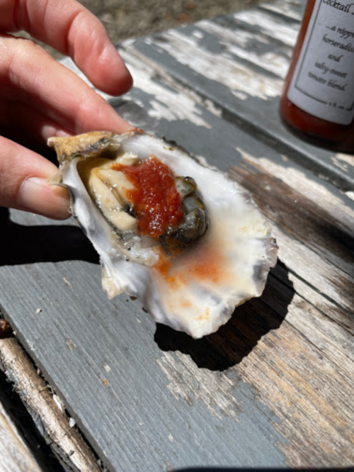 A hand holding an open oyster topped with sauce, resting on a weathered wooden table.