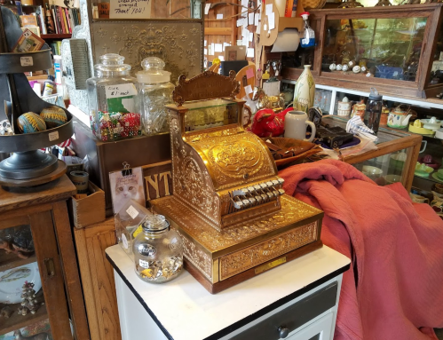 A vintage cash register sits on a table surrounded by various antiques and collectibles in a cozy shop.