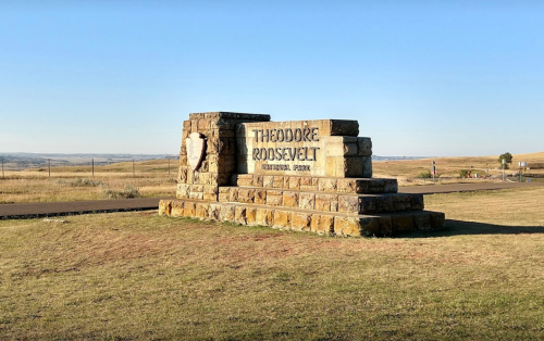 Sign for Theodore Roosevelt National Park, featuring stone structure and grassy landscape under a clear blue sky.