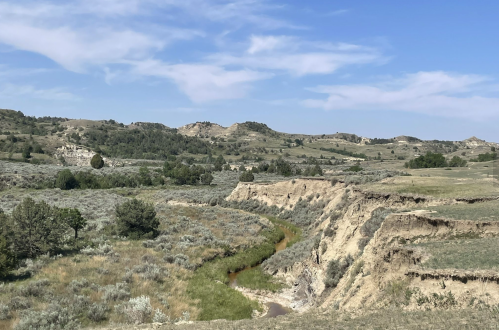 A scenic landscape featuring rolling hills, a winding creek, and sparse vegetation under a blue sky with wispy clouds.