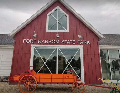 Red barn-style building with "Fort Ransom State Park" sign and an orange wagon in front. Overcast sky.