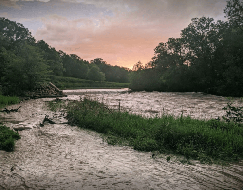 A serene river flows through lush greenery under a cloudy sky at sunset, reflecting soft hues of orange and pink.