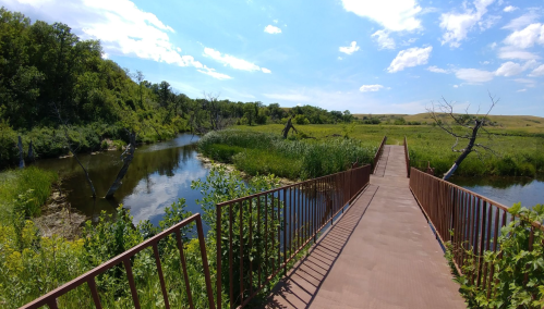 A wooden bridge leads over a calm river, surrounded by lush greenery and blue skies with scattered clouds.