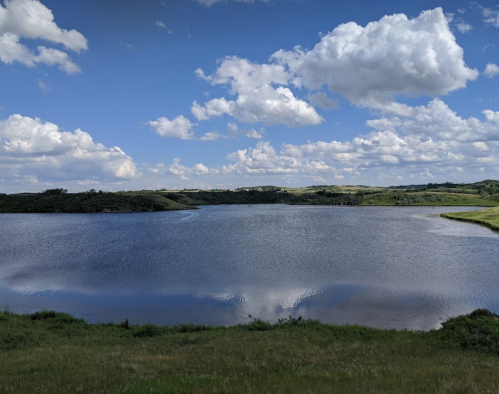 A serene lake surrounded by green hills under a blue sky with fluffy white clouds.