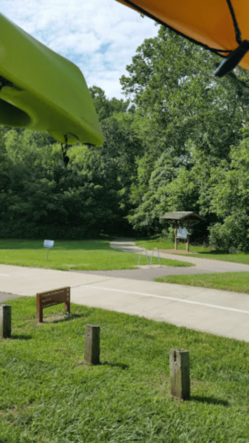 A view of a park path with green grass, trees, and a signpost, partially shaded by colorful kayak canopies.