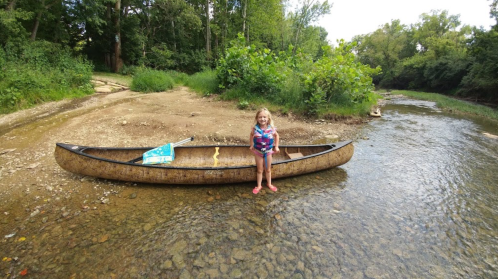 A young girl stands beside a canoe on a sandy riverbank, surrounded by greenery and shallow water.