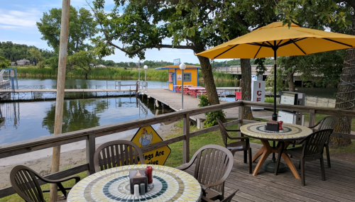 A lakeside view featuring a wooden deck with tables, an umbrella, and a dock with colorful buildings in the background.