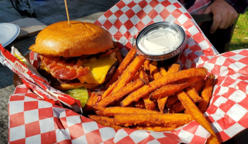 A burger with bacon and cheese, served with sweet potato fries and a side of dipping sauce in a checkered basket.