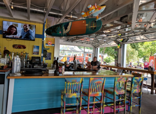 A colorful bar with a surfboard overhead, featuring a TV and patrons enjoying the vibrant atmosphere.
