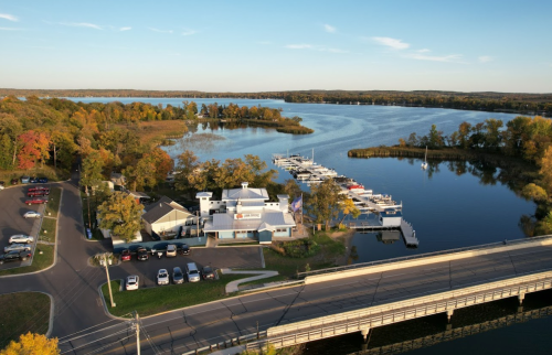 Aerial view of a marina with boats, surrounded by autumn trees and a calm river, near a road and bridge.