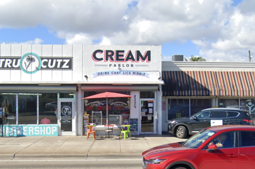 A storefront labeled "Cream Parlor" with outdoor seating and a red umbrella, next to a barbershop.
