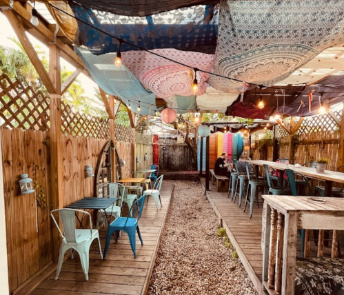 A cozy outdoor seating area with colorful fabric canopies, string lights, and wooden tables along a gravel path.