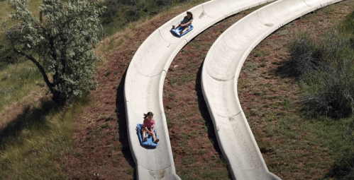 Two children slide down a winding concrete slide on blue sleds, surrounded by grassy terrain and trees.