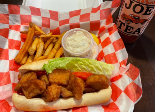 A sandwich with fried chicken, lettuce, and tomato, served with fries and a cup of dipping sauce, alongside a drink.