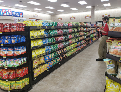 A grocery store aisle filled with various snack chips, with a staff member organizing products in the background.