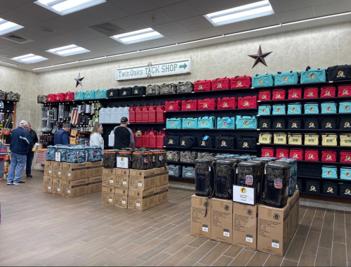 A store interior featuring shelves of colorful bags and boxes, with customers browsing in the Twin Oaks Tack Shop.