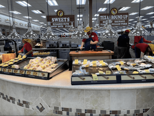 A bustling food counter with staff serving sweets, nuts, and BBQ items in a well-lit store.