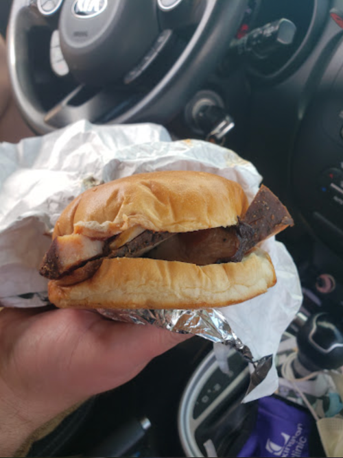 A hand holding a sandwich with meat, wrapped in foil, inside a car. The steering wheel is visible in the background.