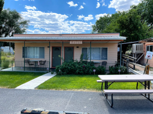 A small, single-story building with two doors, surrounded by grass and trees, featuring outdoor seating.