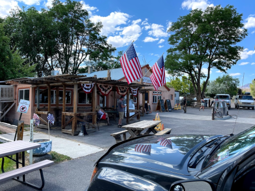 A rustic building with American flags, picnic tables, and a clear blue sky, surrounded by trees and parked vehicles.