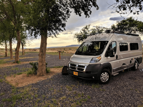 A silver van parked among trees at a scenic campsite, with a cloudy sky and open landscape in the background.