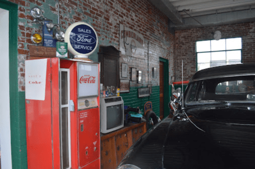 A vintage garage with a classic car, a Coca-Cola vending machine, and retro Ford service signs on the brick walls.