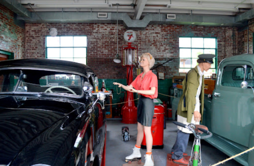 A vintage garage scene featuring a classic car, a mannequin in retro attire, and a man in uniform.