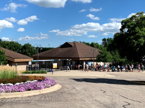 A building with a brown roof surrounded by greenery and flowers, with a line of people outside on a sunny day.